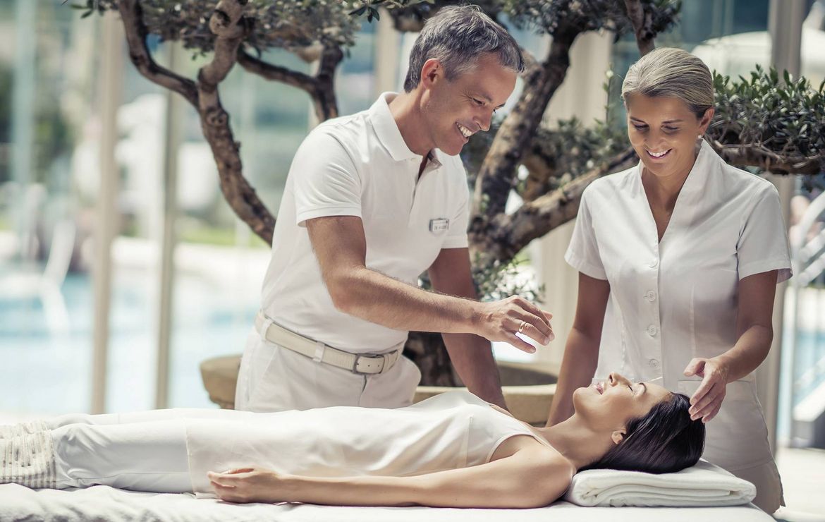 Eine Frau liegt bei einer Medical-Wellness-Behandlung auf der Liege, während zwei Mitarbeiter sie im Spa betreuen.