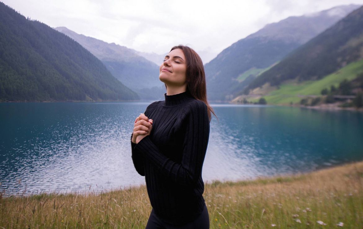 Una donna in nero sta in riva a un lago di montagna con gli occhi chiusi e le mani intrecciate.