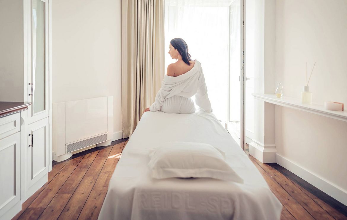 Eine Frau sitzt auf einer Massageliege in einem hellen Behandlungsraum mit Holzfußboden und Blick zum Fenster.