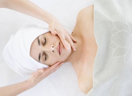 Una donna riceve un trattamento viso spa sdraiata su un lettino bianco con un asciugamano avvolto nei capelli.