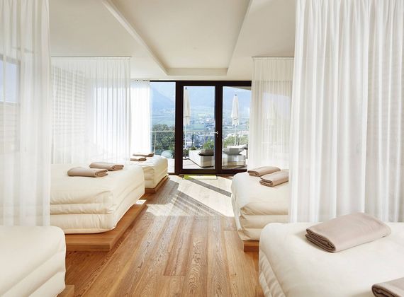 Ein heller Behandlungsraum im Wellnesshotel mit mehreren Liegen, weißen Vorhängen und Blick durch die Fenster auf die Berge.