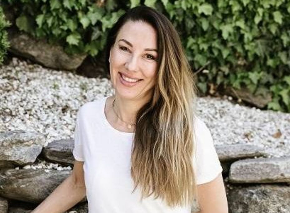 Porträt einer Frau im Preidlhof-Garten Eine lächelnde Frau in weißem T-Shirt sitzt vor einer Steinmauer mit grünen Pflanzen im Hintergrund.