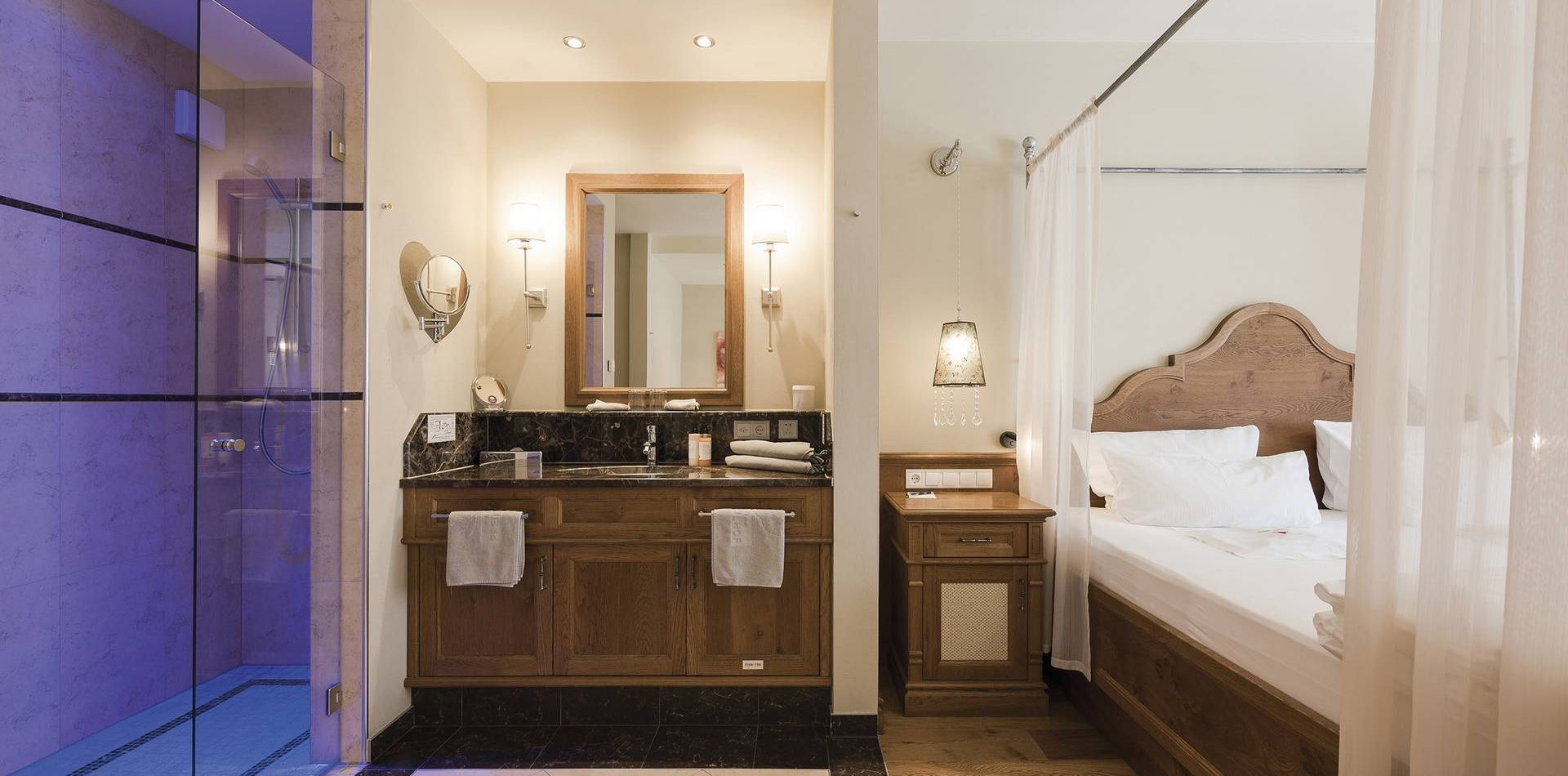 Una suite di lusso con doccia in vetro, doppio lavabo e letto con testiera in legno accanto a una tenda leggera.