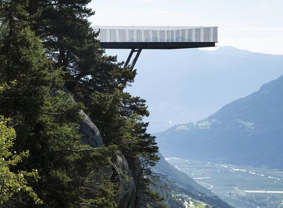 Eine Aussichtsplattform ragt über den bewaldeten Hang und blickt ins weite Tal mit Bergen im Hintergrund.