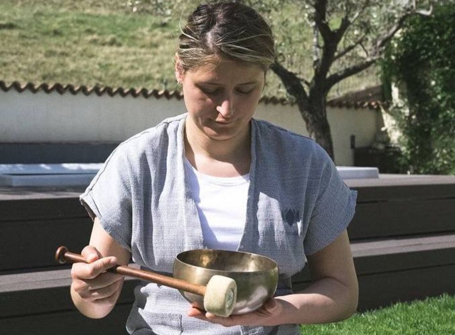 Klangschale im Wellnessbereich des Preidlhof Eine Frau hält im Freien eine Klangschale mit Schlägel in den Händen und sitzt im Garten auf einer Liege.