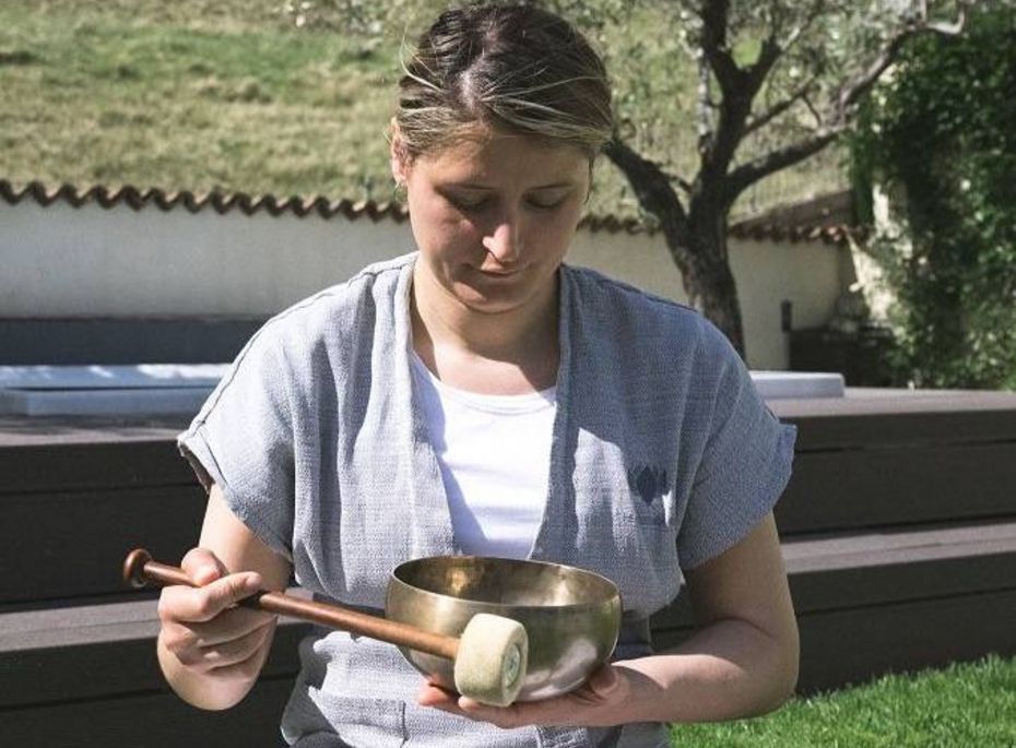 Klangschale im Wellnessbereich des Preidlhof Eine Frau hält im Freien eine Klangschale mit Schlägel in den Händen und sitzt im Garten auf einer Liege.