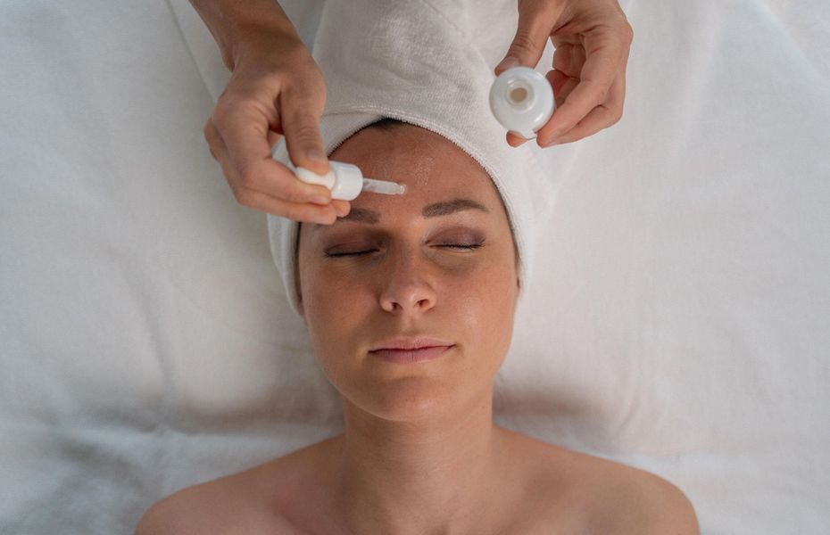 Eine Frau erhält eine Gesichtsbehandlung mit Serum im Spa, während ihr Haar in ein Handtuch gewickelt ist.