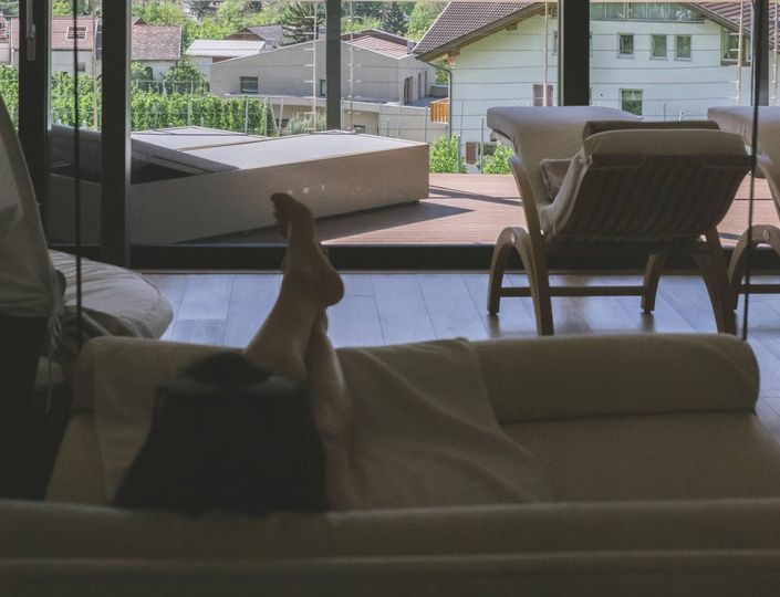 A guest relaxes on a lounge bed beside a window with outdoor deck chairs and garden views.