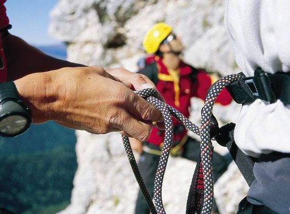 Eine Hand hält ein Kletterseil am Gurt vor einer alpinen Felswand.