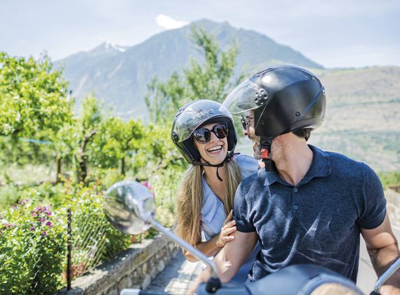 Ein Paar mit Helmen sitzt auf einer Vespa und lächelt vor Bergkulisse im Grünen.