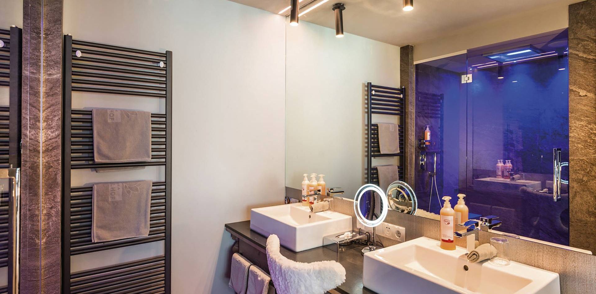 A modern bathroom with two sinks, heated towel rails, a backlit mirror and a glass shower area.