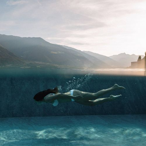 Eine Frau schwimmt im Infinity Pool mit Blick auf die Berge und das warme Licht der tief stehenden Sonne.