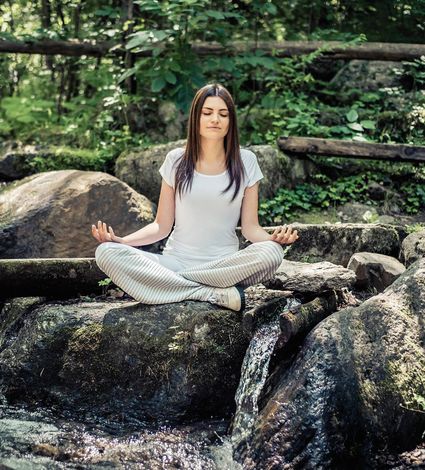 Eine Frau sitzt meditierend im Lotussitz auf einem Felsen an einem kleinen Wasserlauf im Grünen.