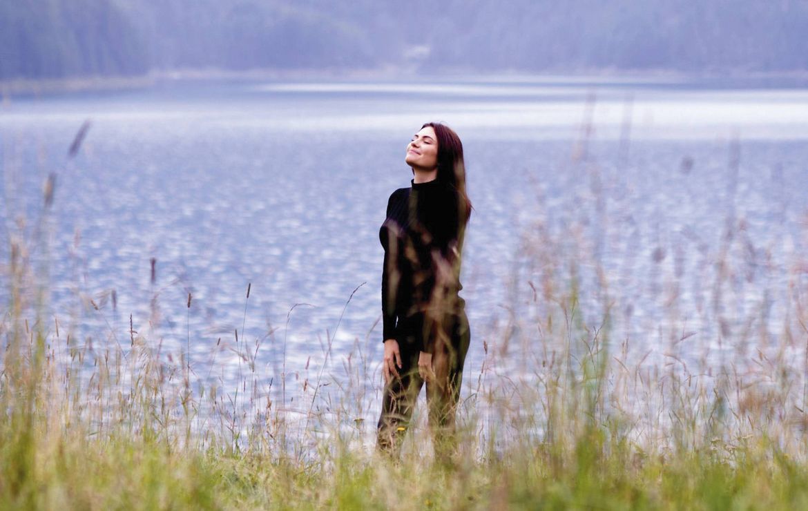 Eine Frau steht entspannt am Ufer vor einem ruhigen See, im Vordergrund hohe Gräser.