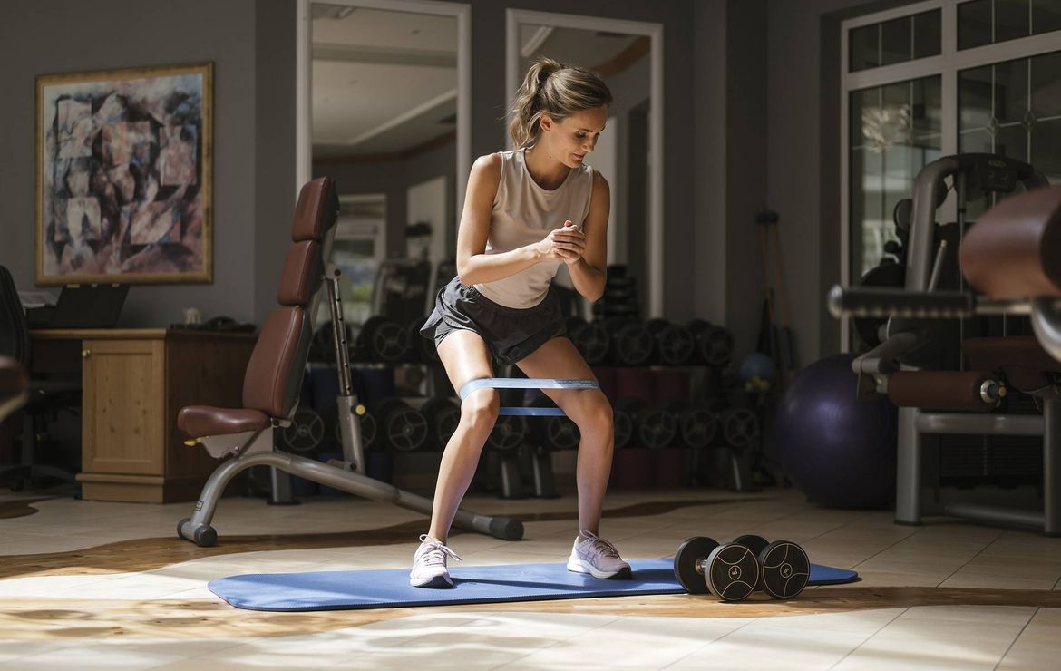 Eine Frau trainiert im Fitnessraum mit einem Widerstandsband und Hanteln auf der Matte.