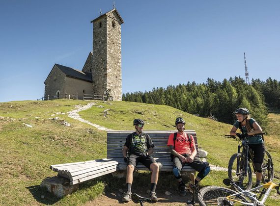Drei Mountainbiker machen Pause auf einer Bank vor einer Bergkapelle auf einer Anhöhe.