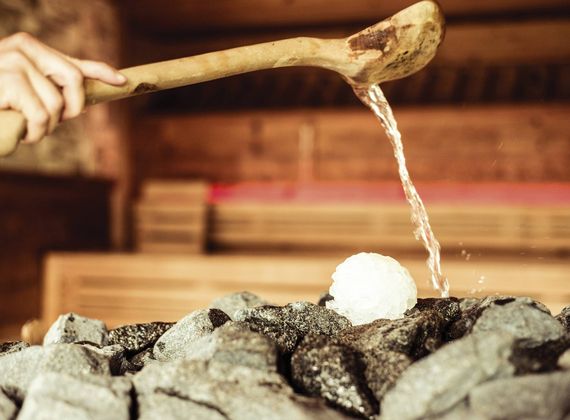 Eine Hand gießt mit einer Holzschöpfkelle Wasser über heiße Saunasteine neben einem weißen Aufgussball.