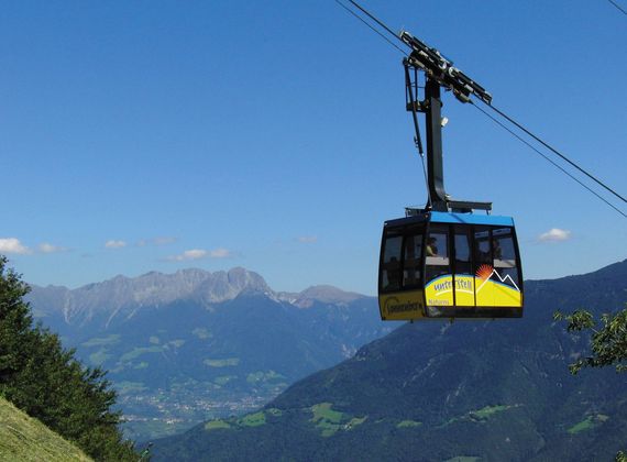 Eine gelbe Seilbahnkabine fährt vor blauem Himmel über das Tal und die Berge bei Naturns.
