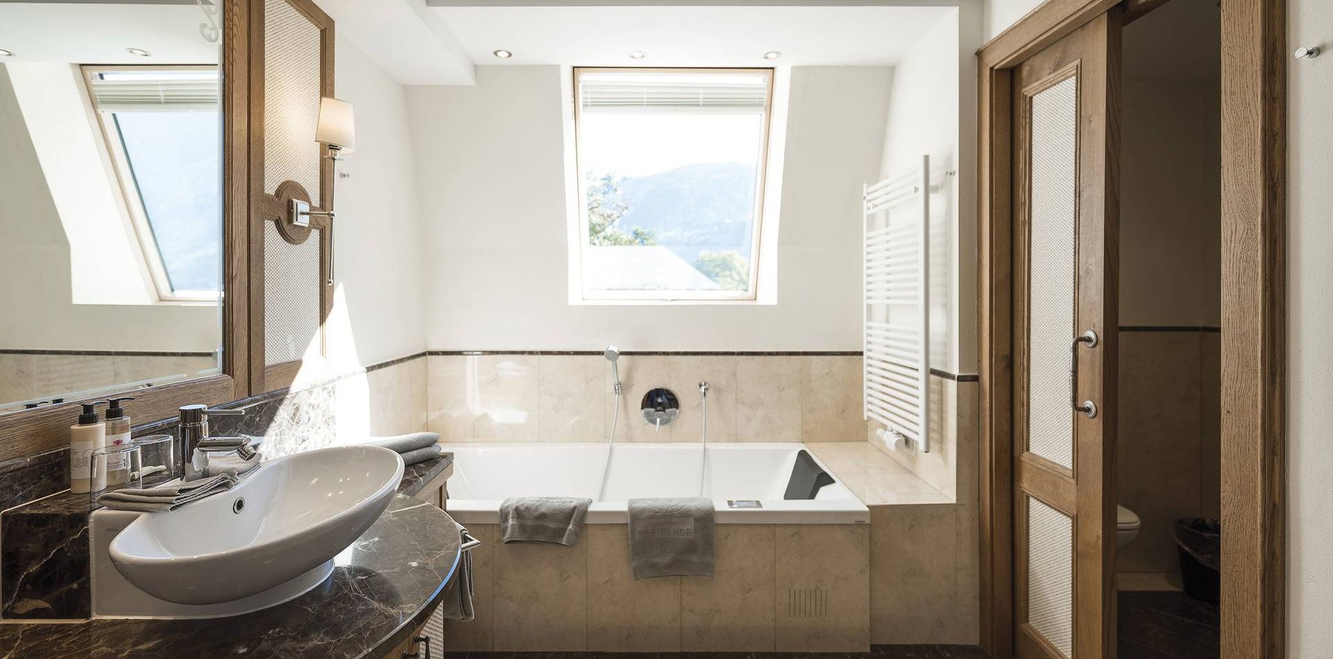 Ein helles Badezimmer mit freistehender Wanne, Waschbecken auf dunkler Ablage und Blick durch das Fenster auf die Berge.