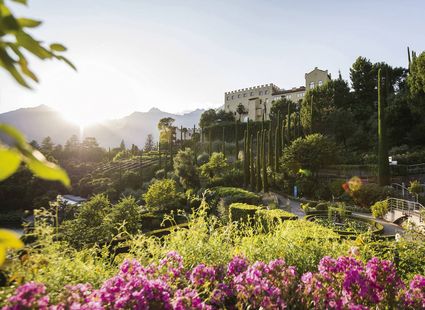 Eine üppige Gartenanlage mit Blüten, Zypressen und dem Hotel auf dem Hügel im warmen Abendlicht.