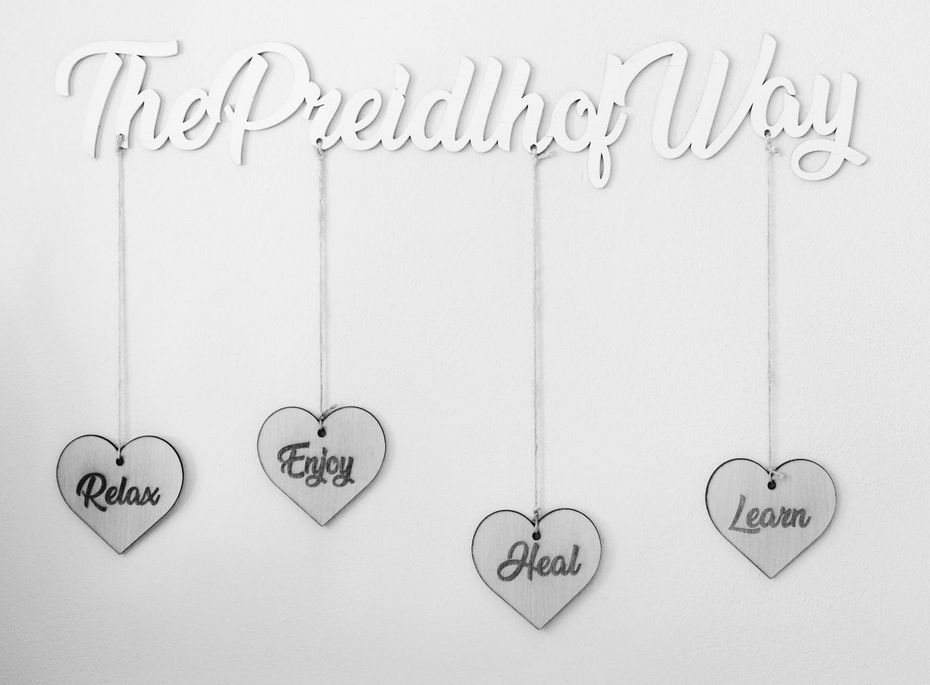 The Preidlhof Way Wanddeko mit Hängeschildern Die Wanddekoration mit der Aufschrift „The Preidlhof Way“ zeigt vier hängende Herzschilder mit den Worten Relax, Enjoy, Heal und Learn.