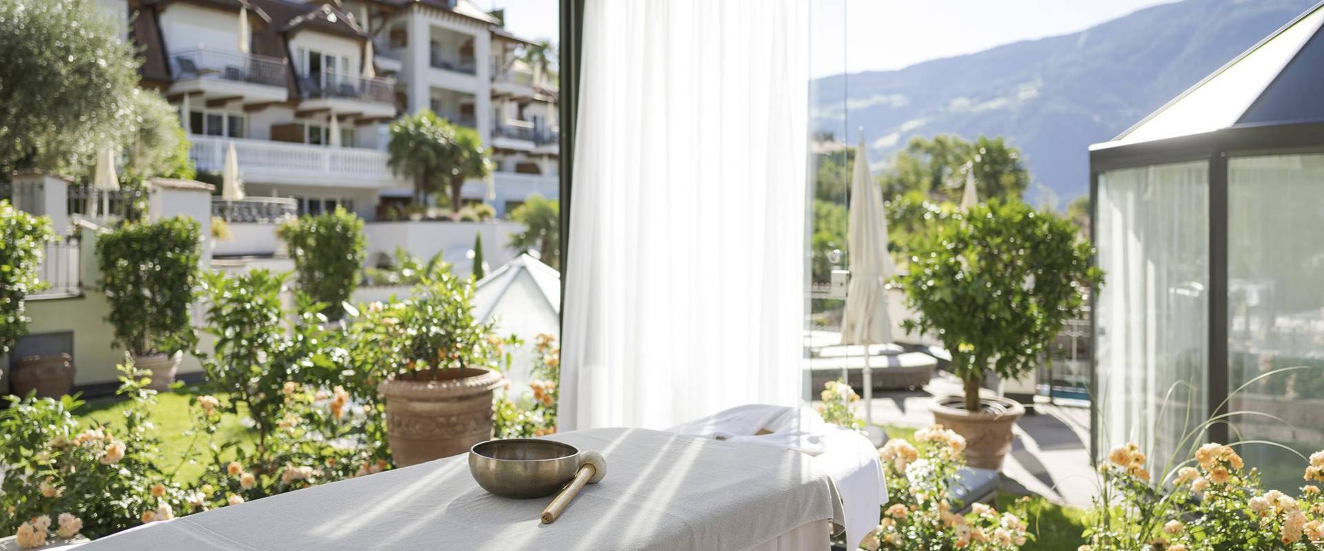 Un lettino da trattamento con una ciotola e un pennello si trova su una terrazza soleggiata con piante in vaso e vista sulle montagne.