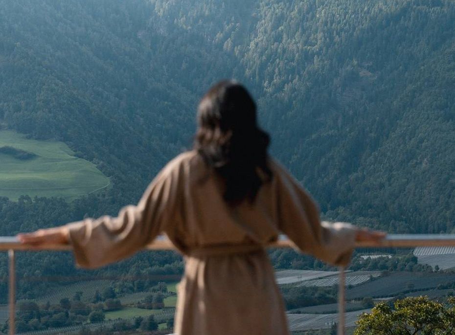 Eine Frau im Bademantel steht auf dem Balkon und blickt über das Tal und die Berge.