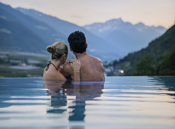 Ein Paar sitzt eng umschlungen im Infinity-Pool mit Blick auf die Berge bei Sonnenuntergang.