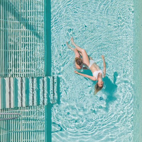 Zwei Personen liegen entspannt im klaren Wasser des Pools neben einer Holzterrasse mit Sonnenliegen.