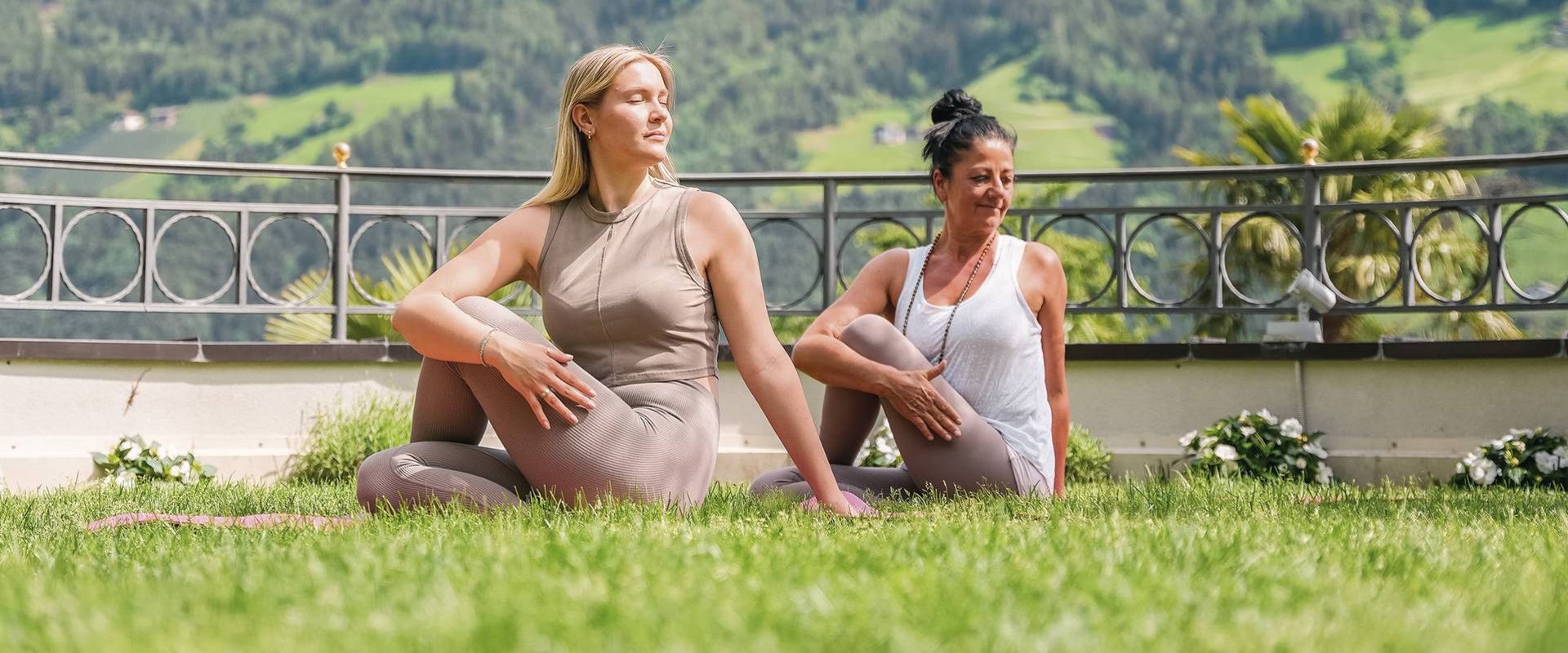 Zwei Frauen sitzen im Gras und üben Yoga mit Blick auf die Berge.