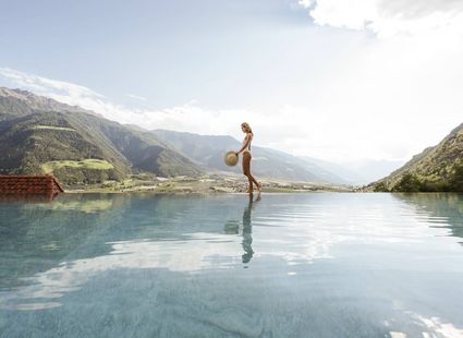 Eine Frau steht am Rand eines Infinity-Pools mit Blick auf die Berge und hält einen Hut in der Hand.