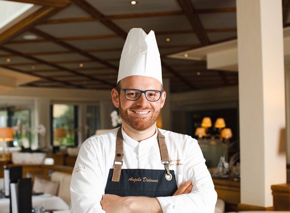 Ein lächelnder Küchenchef mit hoher Kochmütze und Schürze steht im Restaurant mit verschränkten Armen.
