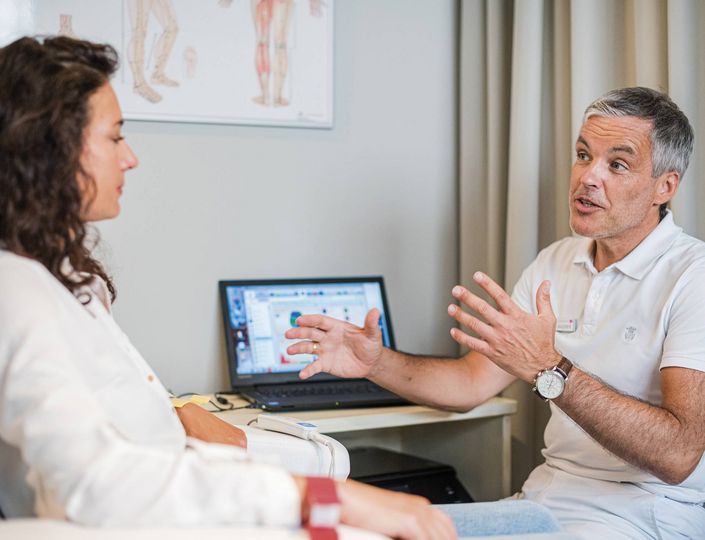 Ein Arzt spricht mit einer Patientin in einem Beratungsraum mit Laptop und Anatomieposter.