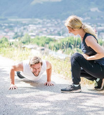 A man does push-ups on a path while a woman crouches nearby and watches during an outdoor fitness session.