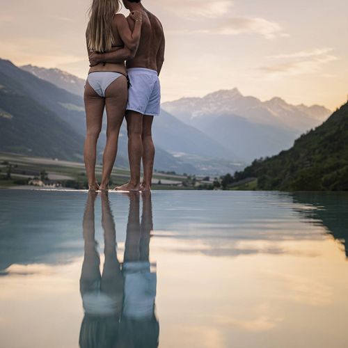 Ein Paar steht im Infinity-Pool und blickt über das Tal in die Berglandschaft bei Sonnenuntergang.