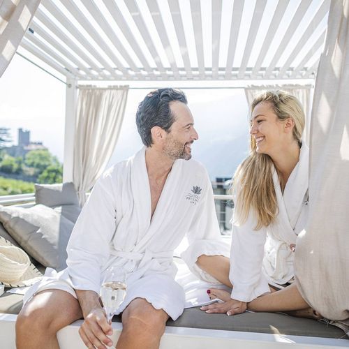 Ein Paar sitzt in weißen Bademänteln in einer hellen Spa-Lounge unter einer Pergola und lächelt sich an.