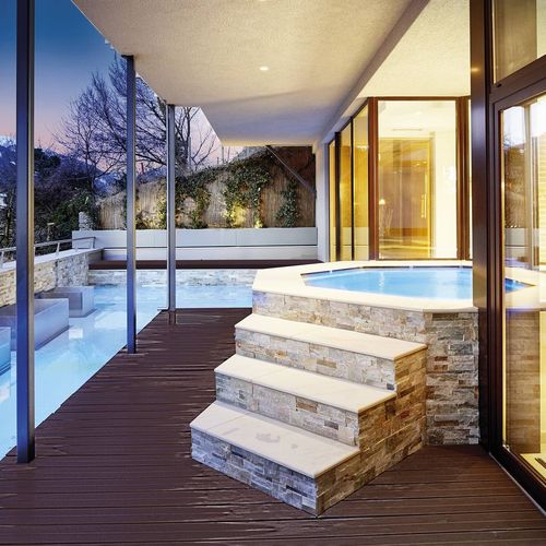 Ein beleuchteter Indoor- und Außenpool mit Holzsteg und Glasfront am Abend.