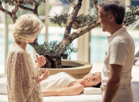 A woman lies on a treatment bed while a couple stands nearby in a bright spa room with an indoor tree.