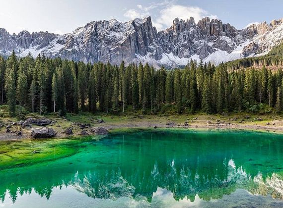 Un lago alpino limpido riflette cime innevate e una fitta foresta di pini sulla riva.