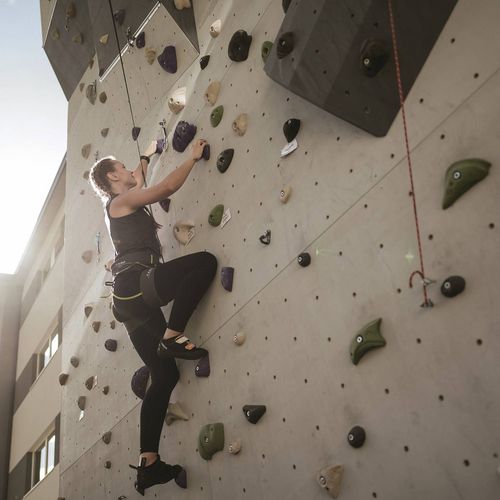 Eine Frau klettert an einer hohen Kletterwand im Freien bei hellem Sonnenlicht.
