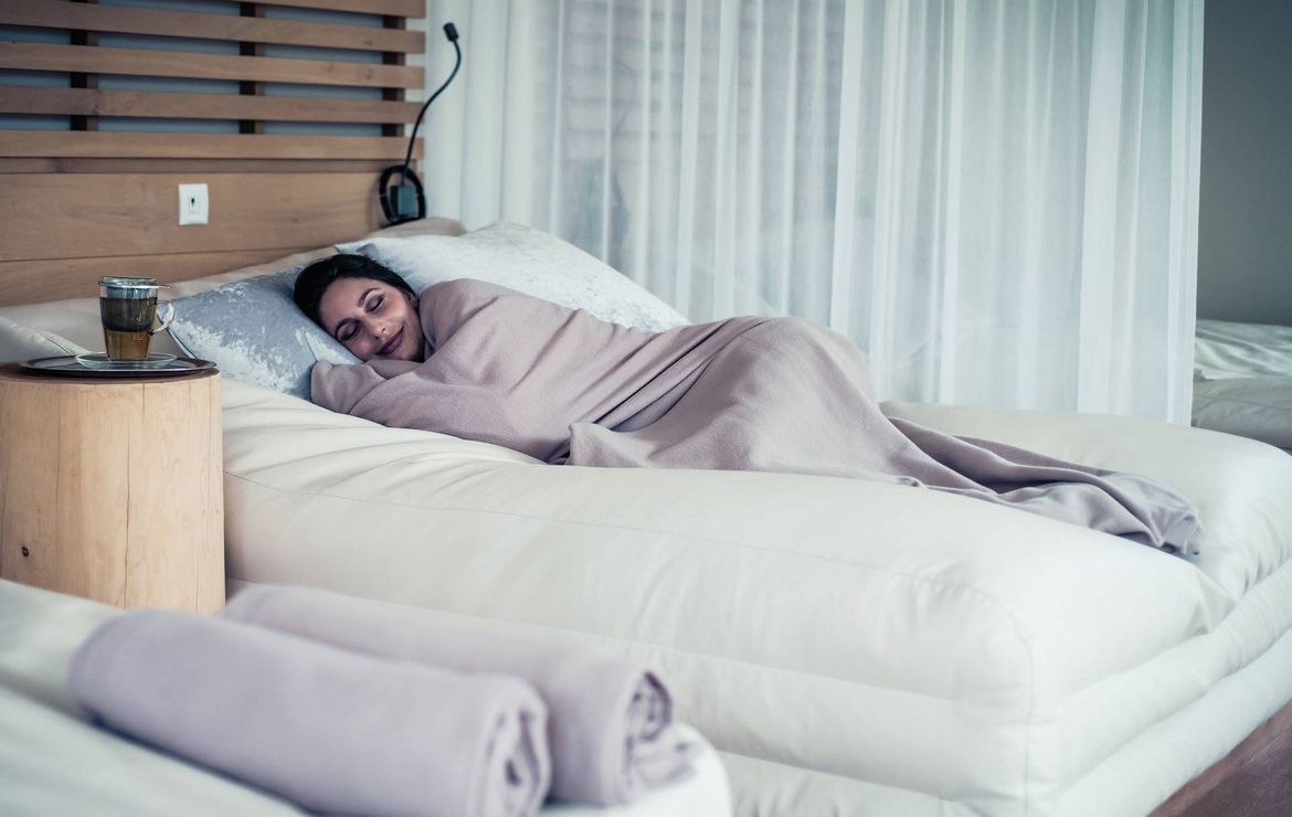 Una donna dorme in un letto ben rifatto con biancheria morbida, tende e un tavolino in una camera tranquilla.