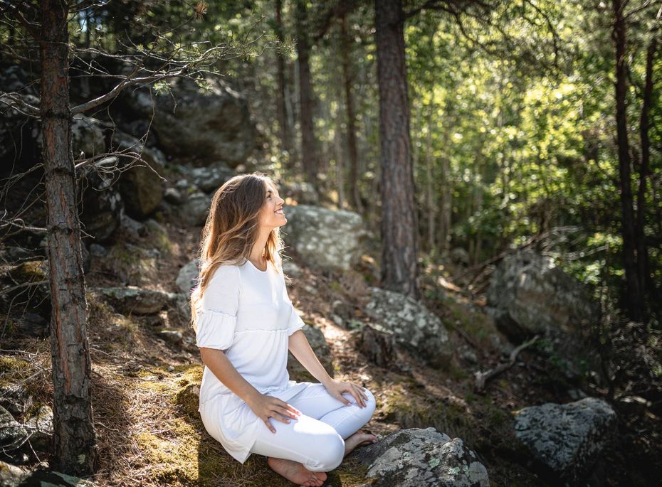 Eine Frau sitzt in Weiß auf einem Felsen im Wald und meditiert in ruhiger Natur.