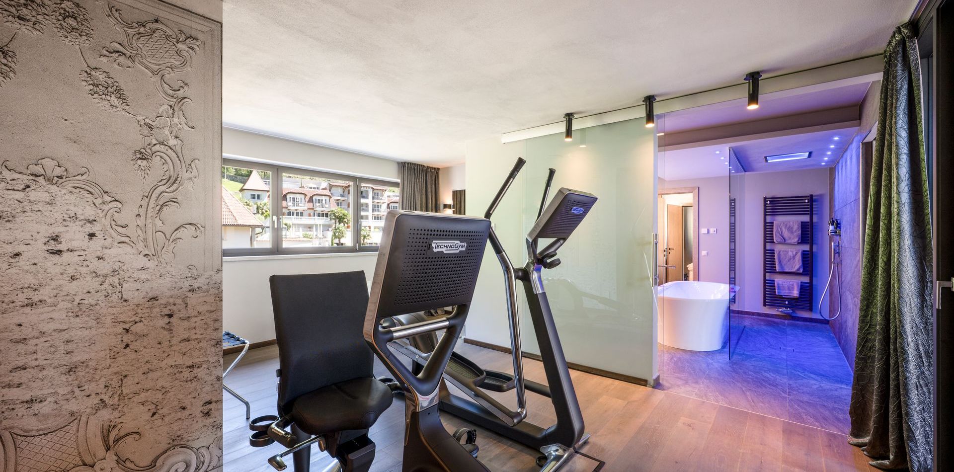 Ein Fitnessbereich in der Penthouse Suite mit Crosstrainer, Stuhl, Badewanne und blau beleuchtetem Bad.
