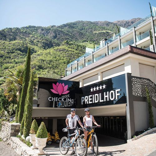 Zwei Radfahrer stehen vor dem Eingang des Preidlhof Wellnesshotels mit Bergkulisse.