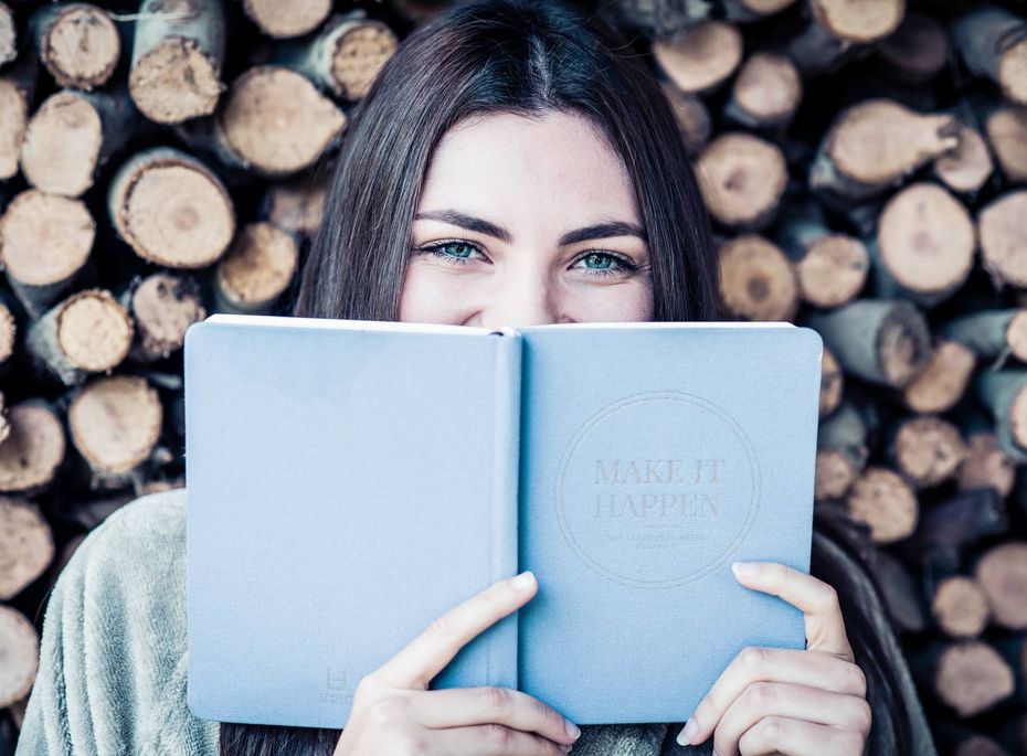 Frau liest im Preidlhof Blog vor Holzstapel Eine Frau hält ein hellblaues Notizbuch vor das Gesicht und blickt über den Rand, vor einem Stapel Holzscheite.