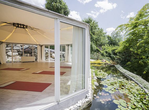 A glass-walled spa pavilion stands beside a pond with lily pads and greenery.