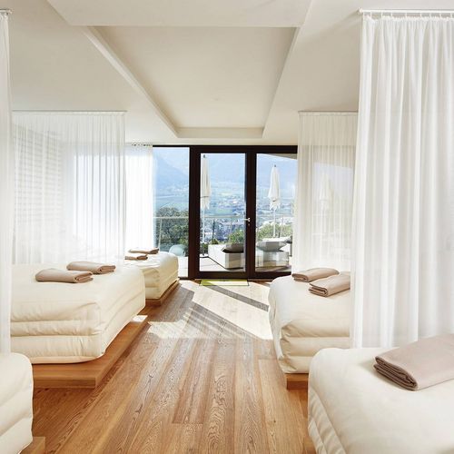 Ein heller Ruheraum im Spa mit mehreren Liegen, Vorhängen und Blick durch die Glasfront nach draußen.