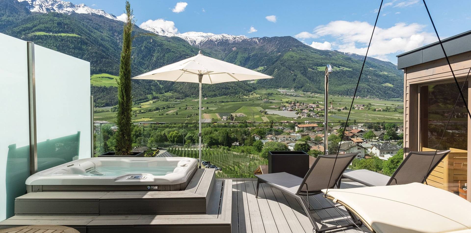 A penthouse suite terrace with a jacuzzi, lounge chairs, an umbrella and mountain views.