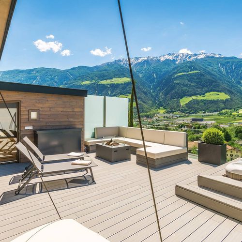 Eine sonnige Hotelterrasse mit Sitzlounge, Liegen und Whirlpool mit Blick auf das Tal und die Berge.