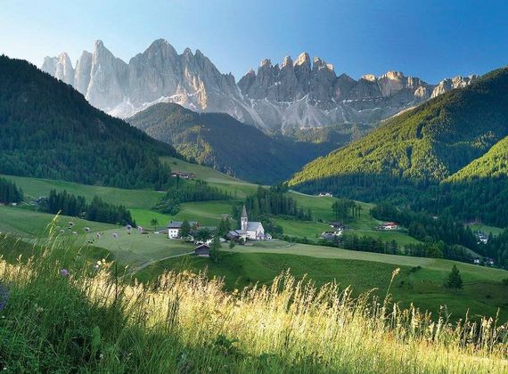 Un villaggio di montagna si trova in una valle verde sotto cime frastagliate e prati alpini assolati.